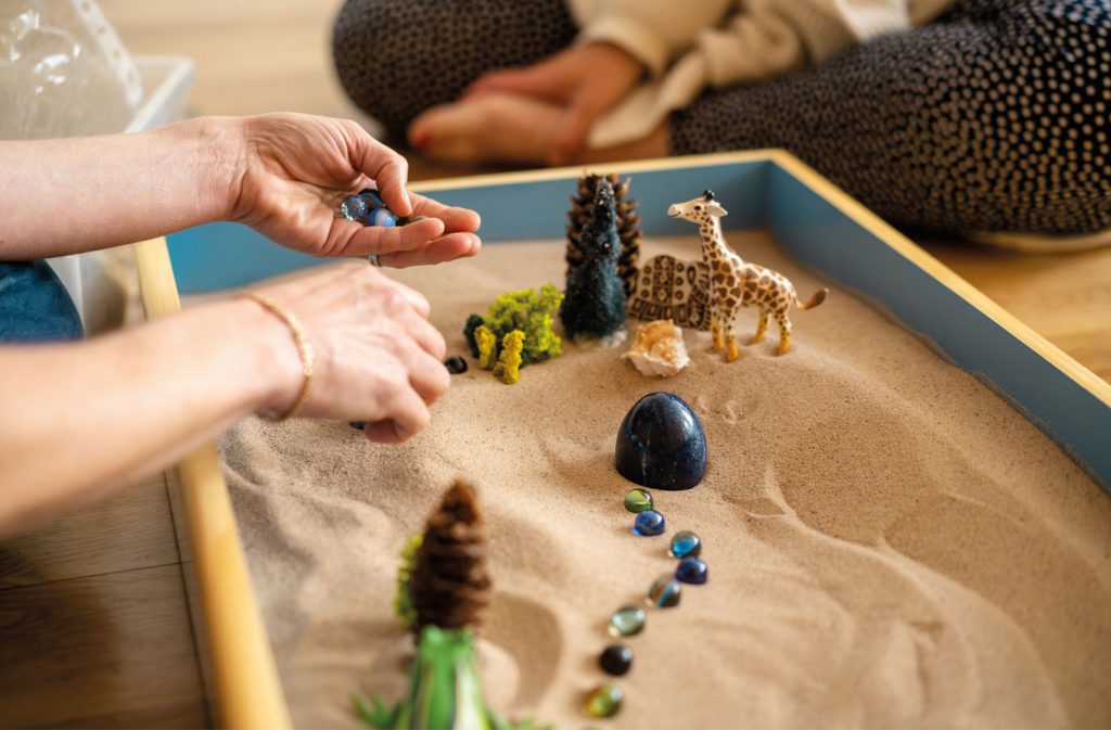 A child arranging miniature figures, a giraffe, trees, and stones, in a sand tray during therapy